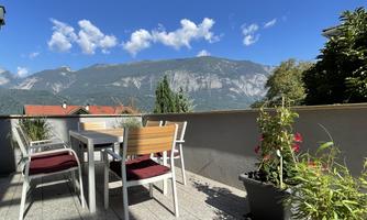 A beautiful balcony with a table and chairs, surrounded by plants. In the background, the mountains and a blue sky can be seen.