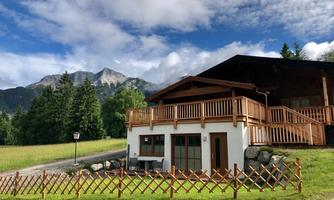 A modern chalet surrounded by green meadows and trees. In the background, majestic mountains can be seen under a partly cloudy sky.