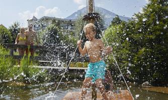 A happy child plays on a wooden raft and splashes with water. In the background, adults and a green environment can be seen.
