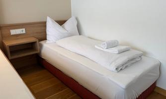 A simple single room with a bed that is prepared with fresh bed linens and towels. Next to the bed is a nightstand.
