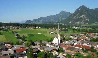 A picturesque view of a village surrounded by green fields and mountains. In the center stands a church with a tall steeple.
