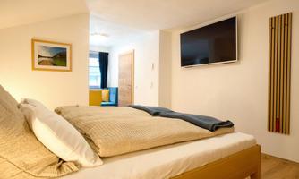 A modern bedroom with a cozy bed and a large TV on the wall. The bright decor creates an inviting atmosphere.