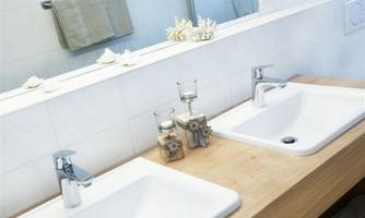 A modern bathroom with two sinks on a wooden shelf. The walls are light and decorated with candles and a towel.