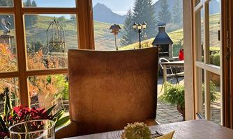 A cozy table with a view of the mountains and nature. In the foreground, flowers and a glass of water are visible.