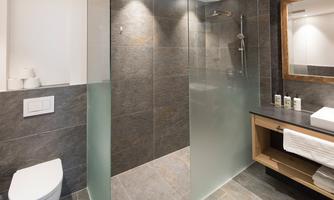A modern bathroom with a glass shower and a stylish wooden shelf. The walls are in neutral gray tones, and there is bright lighting.