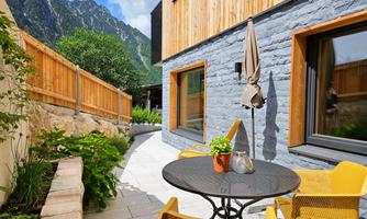 A cozy outdoor area with a table and yellow chairs. In the background, green plants and tall mountains are visible.