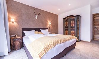 A cozy bedroom with a double bed and a padded wall. In the background, there is a rustic wardrobe, and a deer antler is hanging on the wall.