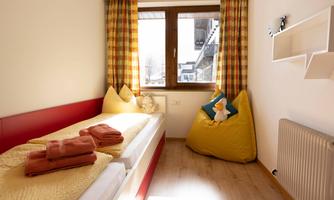 A cozy room with a single bed and colorful cushions. The large windows let in a lot of light and offer a view of the outside world.