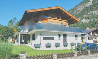 A modern house in Alpine style with wooden details and a balcony. It is situated in a picturesque setting with mountains in the background.
