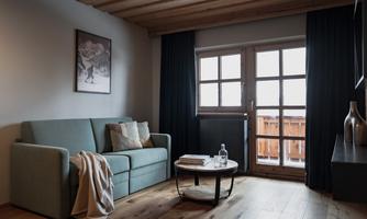 A cozy living room with a light blue sofa and a round table. Large windows bring in daylight and offer a view of the balcony.