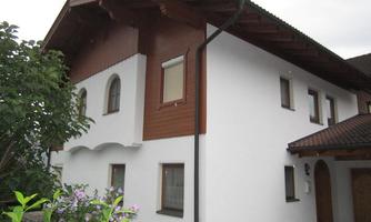 A modern house with white and wooden cladding. The surroundings are adorned with green plants and flowers.