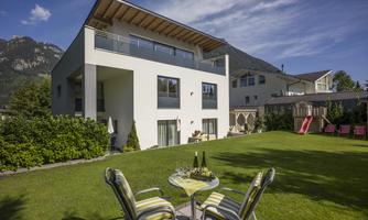A modern house with a spacious garden area. In the foreground, there are two chairs and a small table with drinks.