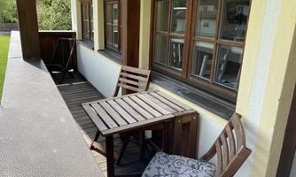 A small balcony with a table and two wooden chairs. The windows offer a view of the outdoors and the weather is sunny.