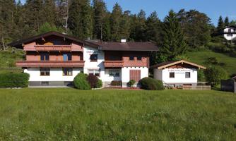 A large, traditional house surrounded by green grass and trees. In the background, more buildings and mountains are visible.