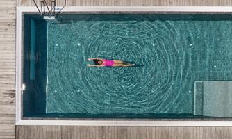 A swimming pool with clear water and a wooden deck. A person in a pink swimsuit is swimming relaxed on the water.