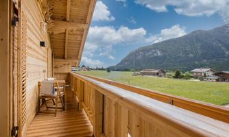 A cozy wooden balcony overlooking green meadows and mountains. The sky is partly cloudy and the surroundings are quiet and picturesque.