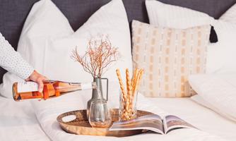 A cozy bed with white pillows and a tray. On the tray, there is a bottle of rosé wine, a vase with dried flowers, some snacks, and a magazine.