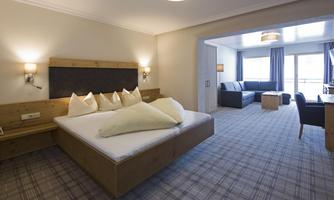 A spacious hotel room with a large bed and fresh bedding. There is a living area with a sofa set and a balcony.