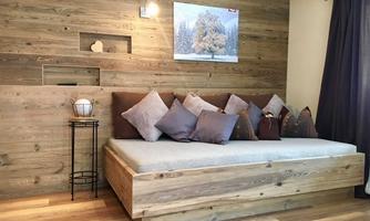 A cozy living room with wood paneling and a low bed. There are several cushions and a small side table.