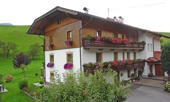 A charming wooden house with colorful flowers on the balconies. It is located in a green landscape with gentle hills.