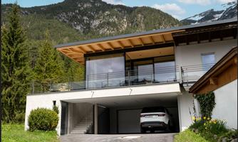 A modern house in the mountains with large windows and a covered parking space. In the background, green trees and snow-capped mountains can be seen.