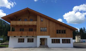 A traditional wooden house with two floors and a balcony. The sky is blue with some white clouds.