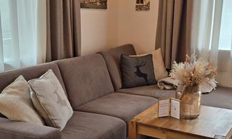 A cozy living room with a beige sofa and decorative cushions. On the table, there is a vase with dried flowers and some small decorative objects.