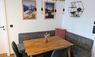 A cozy dining area with a wooden table and a corner bench. Pictures of mountains hang on the wall, and a modern lamp provides light.
