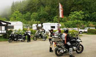 A group of motorcyclists is standing in a parking lot between RVs. In the background, trees and a flag can be seen.