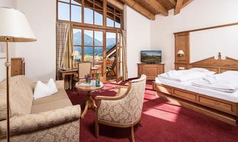 A cozy room with a wooden ceiling, a large window, and a view of the mountains. There is a bed, a sofa area, and a television.