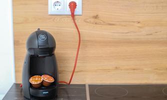 A coffee machine is on a kitchen countertop, with two coffee capsules in the holder. In the background, there is a red electrical outlet on a wooden wall.