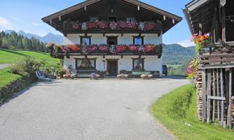 A traditional farmhouse with flowered balconies in a rural setting. In the background, green meadows and mountains can be seen.