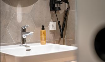 A modern bathroom with a white sink and a stainless steel faucet. On the shelf there is a bottle of shampoo and a hairdryer is mounted on the wall.