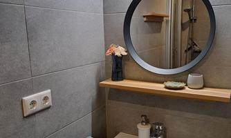 A modern bathroom with gray tiles and a round mirror. On a wooden shelf, there are flowers and bathroom accessories.