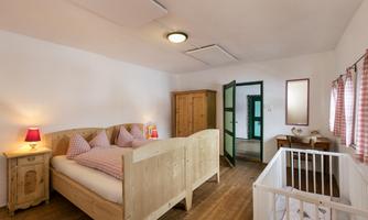 A cozy bedroom with a wooden bed, pillows, and a small crib. There is a desk and a window with curtains.
