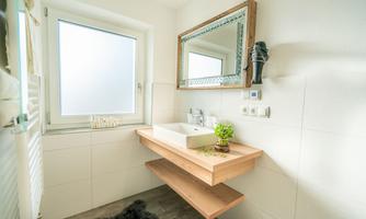 A modern bathroom with a wooden vanity and a large mirror. Light streams in through a window, creating a bright, inviting atmosphere.