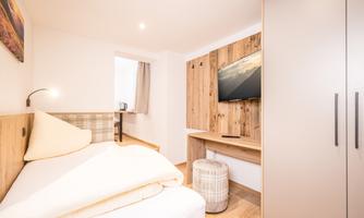 A modern bedroom with a single bed and wood-paneled walls. There is a television, a wardrobe, and pleasant lighting.