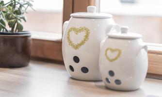 Two white pots with a heart-shaped pattern are on a table. The larger pot has black dots and the smaller pot is simply designed.