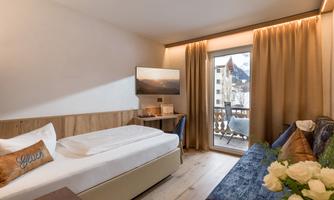 A modern hotel room with a single bed and a cozy seating area. Large windows provide plenty of daylight and offer a view of a balcony.