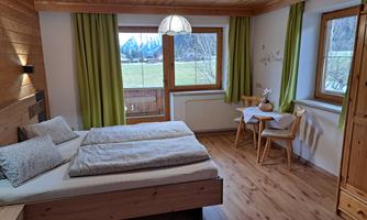 A cozy bedroom with a double bed and wooden furniture. Large windows with green curtains let in plenty of light and offer a view of nature.