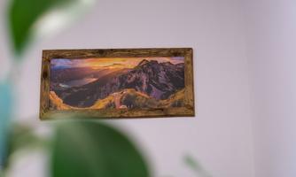 A beautiful landscape painting with mountains and a sunset, framed in a rustic wooden frame. In the foreground, a green plant is visible.