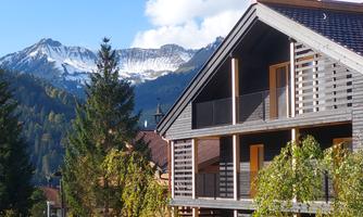 A modern wooden house with a balcony in front of a picturesque mountain landscape. In the background, snow-covered mountains and a blue sky can be seen.