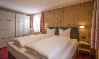 A cozy bedroom with a double bed and two pillows. The walls are made of wood, and there is a white wardrobe as well as windows with curtains.