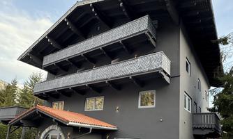 A modern three-story building with a dark paint and several balconies. In front of the entrance is a red roof and a small terrace.