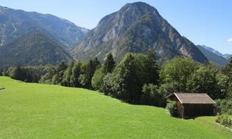 A green meadow with a small wooden house surrounded by mountains. In the background, high mountains are visible under a clear blue sky.