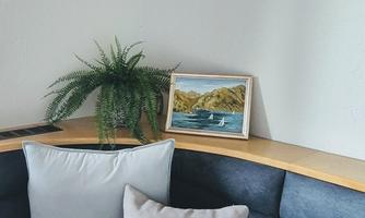 A cozy seating area with cushions and a small table. In the background, there is a plant arrangement and a framed picture of a seascape.