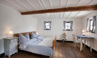 A cozy bedroom with a double bed and light wooden flooring. The room has small windows with checkered curtains and simple furnishings.