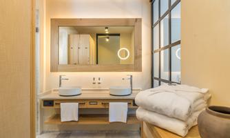 A modern bathroom with wooden shelves, two sinks, and a large mirror. Soft towels are neatly stacked on the shelf.