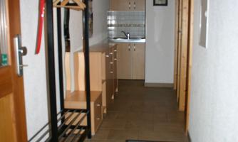 A flat hallway with a coat rack and a small kitchen in the background. The floor is tiled and the area is well lit.