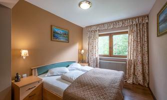 A cozy bedroom with a large double bed and wooden flooring. The windows offer a view of greenery, and curtains with a floral pattern complete the ambiance.